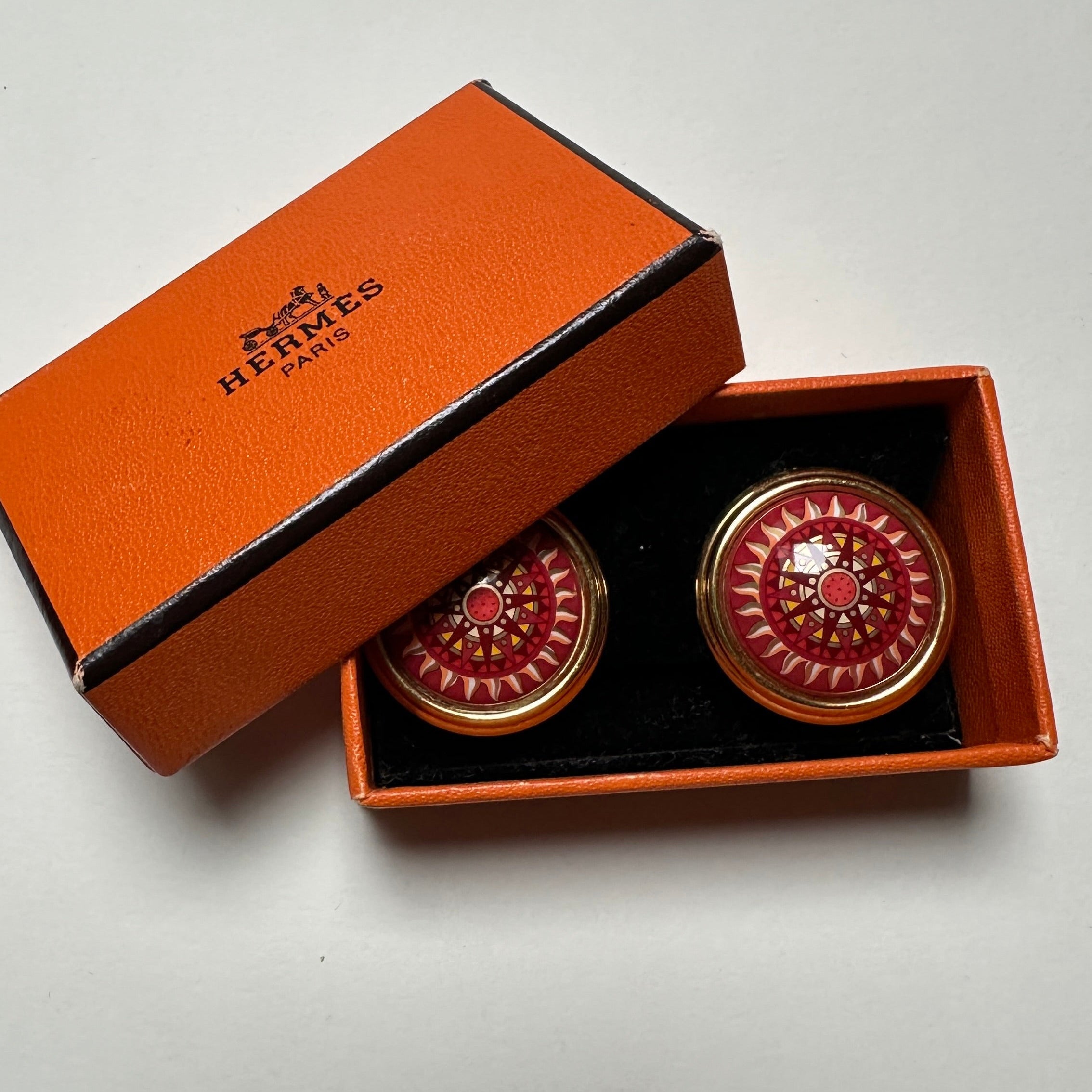 Paire de clips d'oreilles Hermès vintage en émail rouge vif motif baroque doré, présentés dans leur boîte orange d'origine ouverte.