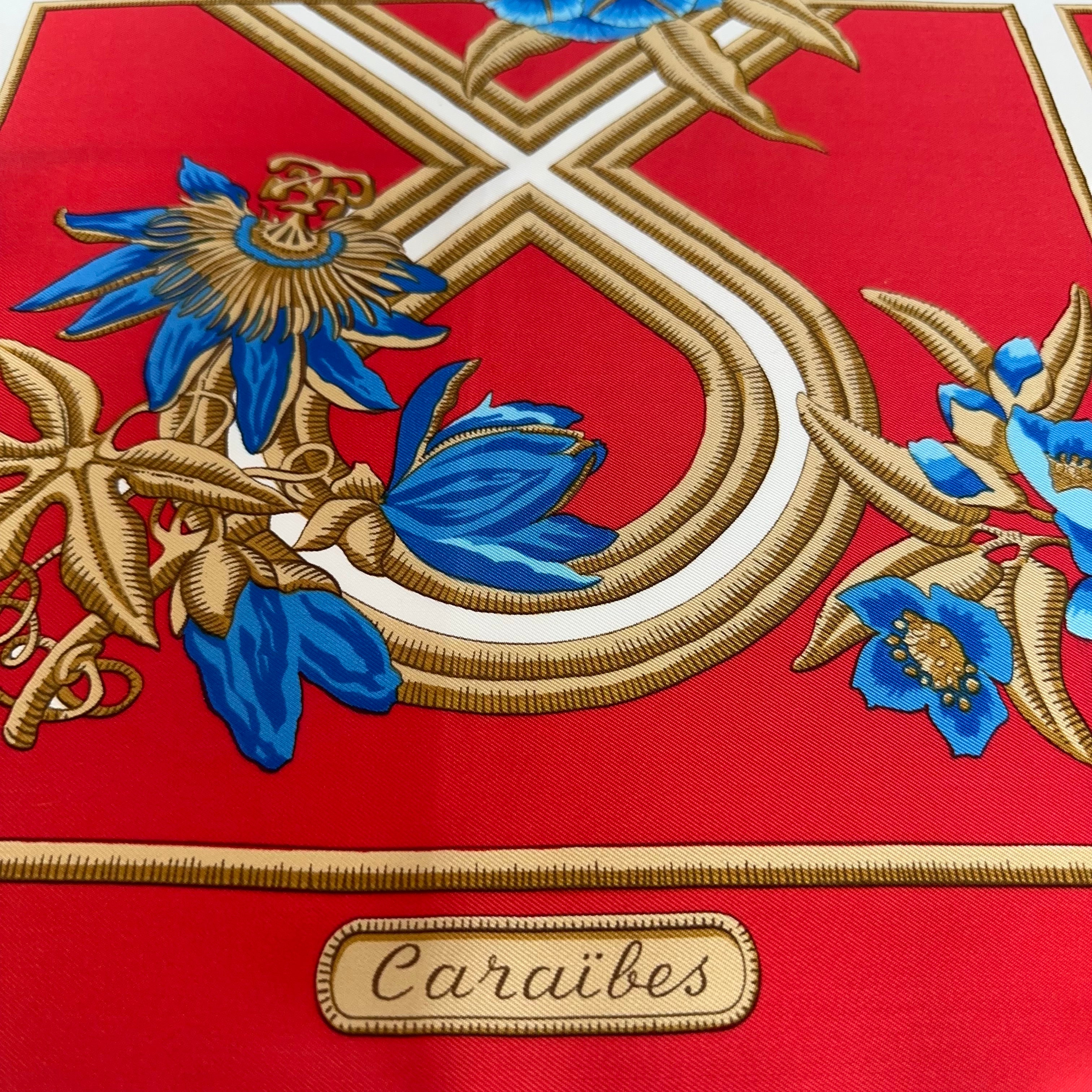 Zoom sur le bas du foulard : fleurs de la passion (passiflores) bleues et or, et cartouche avec le titre "Caraïbes" sur fond rouge.